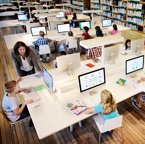 Une adulte enseigne l'utilisation d'ordinateur à des enfants dans une bibliothèque. Chaque enfant est devant un ordinateur mais ils regardent vers la professeure qui parle