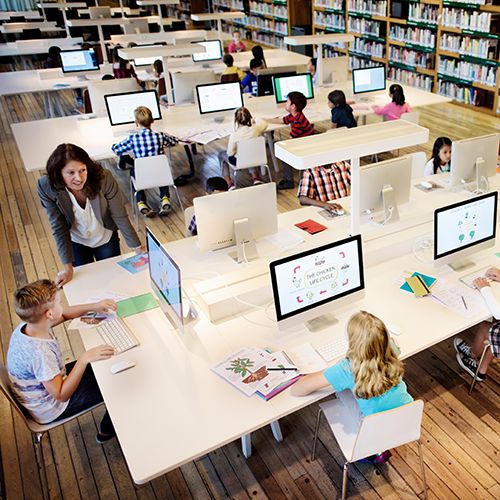 Une adulte enseigne l'utilisation d'ordinateur à des enfants dans une bibliothèque. Chaque enfant est devant un ordinateur mais ils regardent vers la professeure qui parle